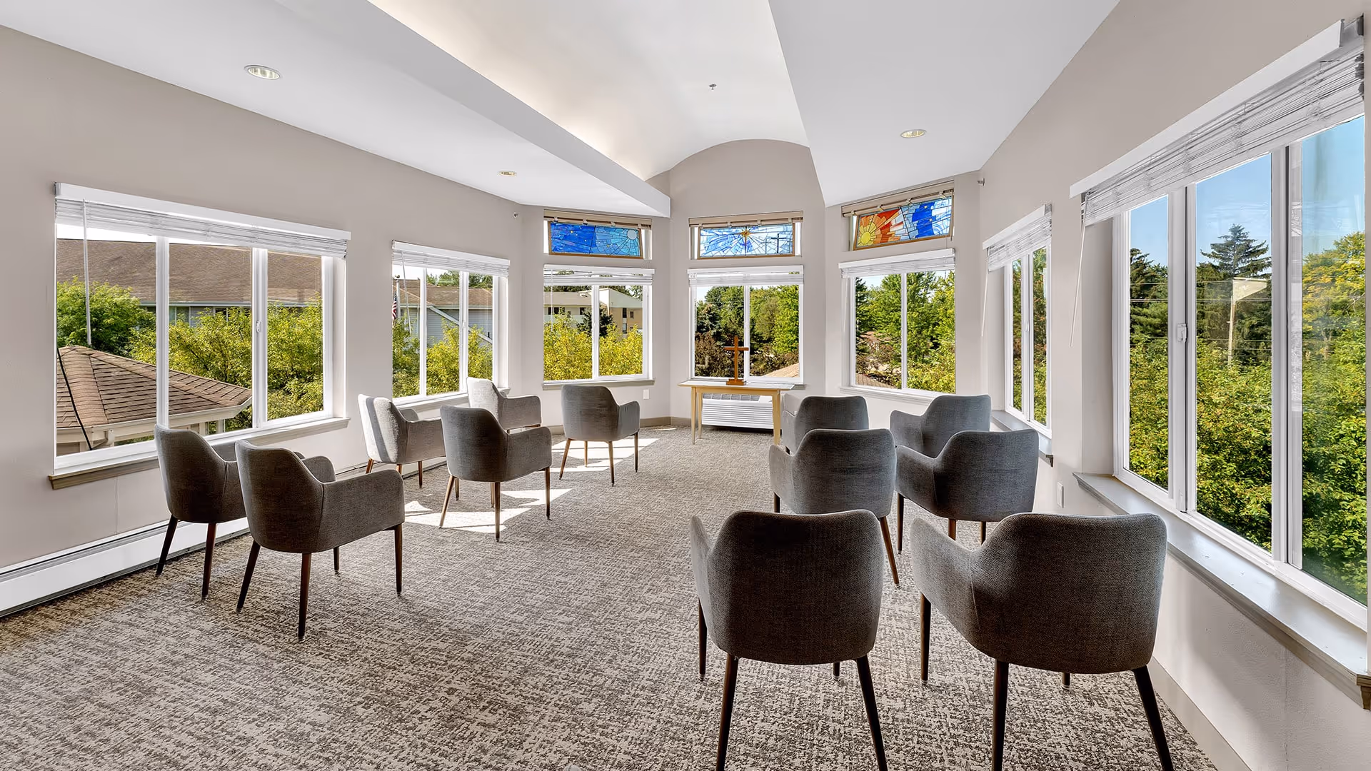 A bright, spacious room with large windows on three sides letting in natural light and offering views of greenery outside. The room contains several modern gray armchairs arranged in a semi-circle facing a small wooden table near the windows. The ceiling is vaulted with recessed lighting, and there are colorful stained glass panels above some windows.