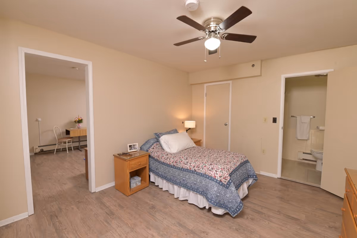 Furnished bedroom with a single bed, nightstand, ceiling fan, and doorways to a bathroom and adjacent room.