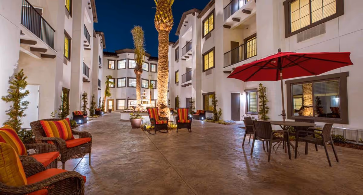 Night view of an outdoor courtyard area at The Park At Laguna Springs featuring wicker chairs with red and yellow cushions, a table with a red umbrella, tall palm trees, potted plants, and a multi-story building with lit windows surrounding the courtyard.