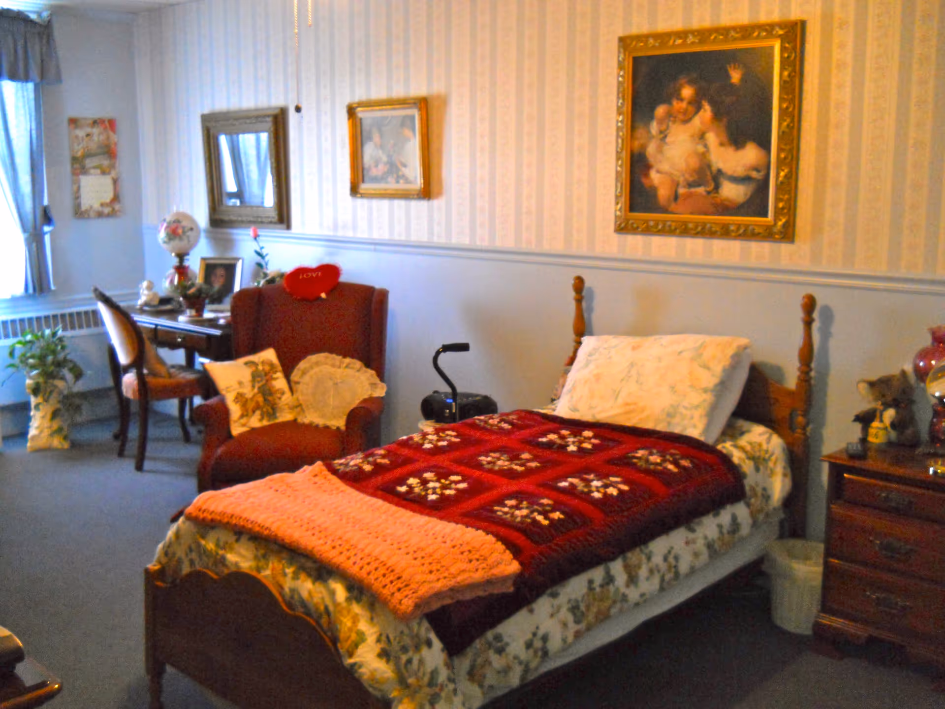 A cozy bedroom with a single wooden bed covered with floral sheets, a red and pink crocheted blanket, and a pillow. Next to the bed is a wooden nightstand with a lamp and a teddy bear. A red armchair with decorative pillows is positioned near a small wooden desk with a chair, a lamp, and framed photos. The walls are decorated with framed paintings and a mirror. A window with curtains allows natural light into the room.