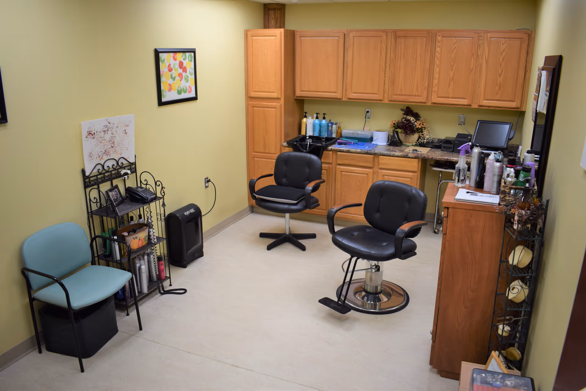 Small hair-styling room with two black salon chairs, cabinets, countertop with hair products, and a waiting chair.