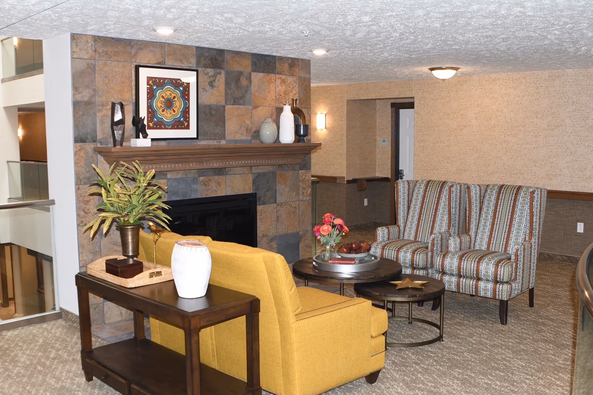 Cozy common seating area with a tiled fireplace, a yellow armchair, two striped accent chairs, and a coffee table with flowers and decor.
