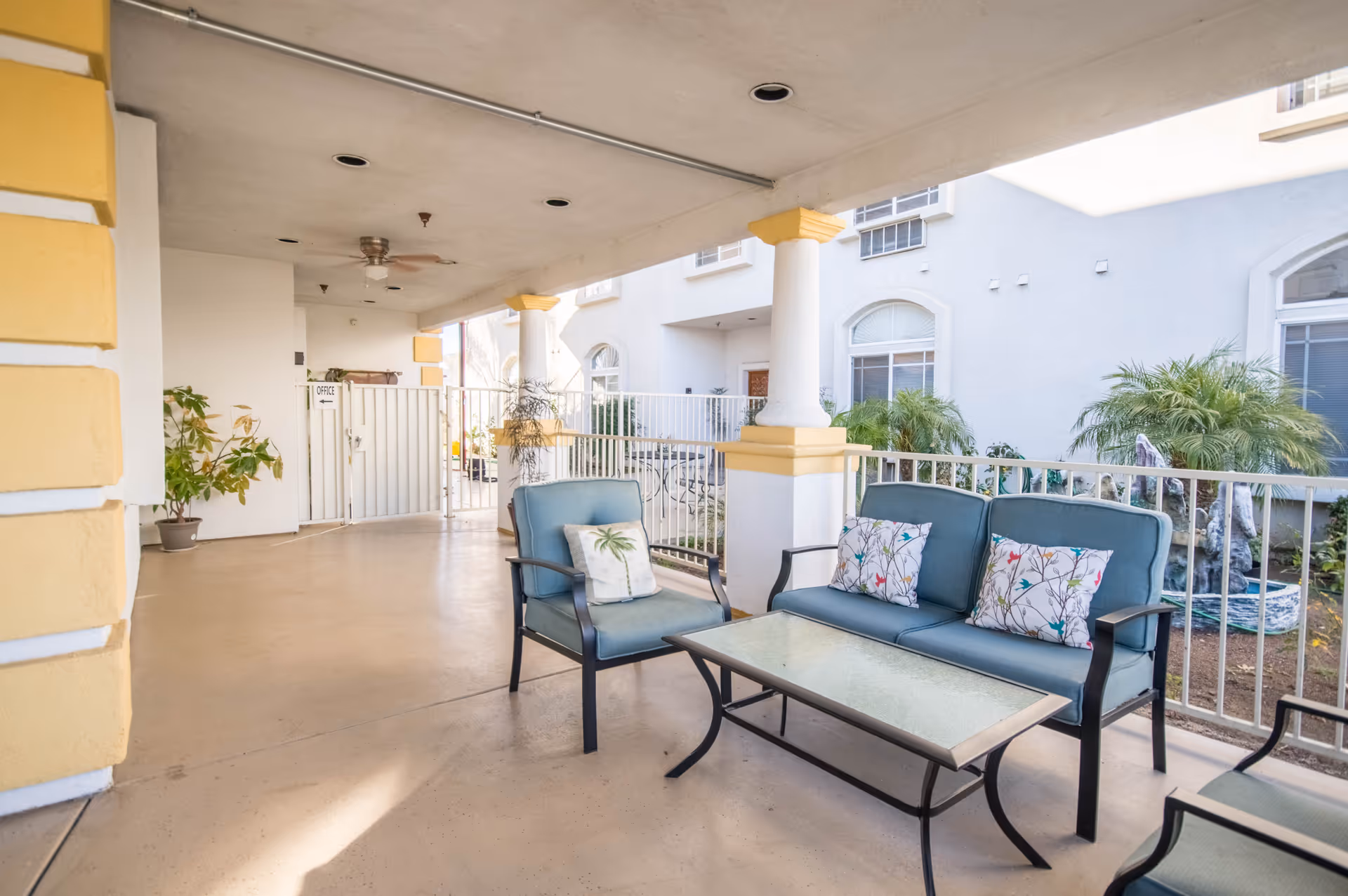 Covered outdoor patio with cushioned chairs and a glass-top coffee table beside a gated entrance to the assisted living building.