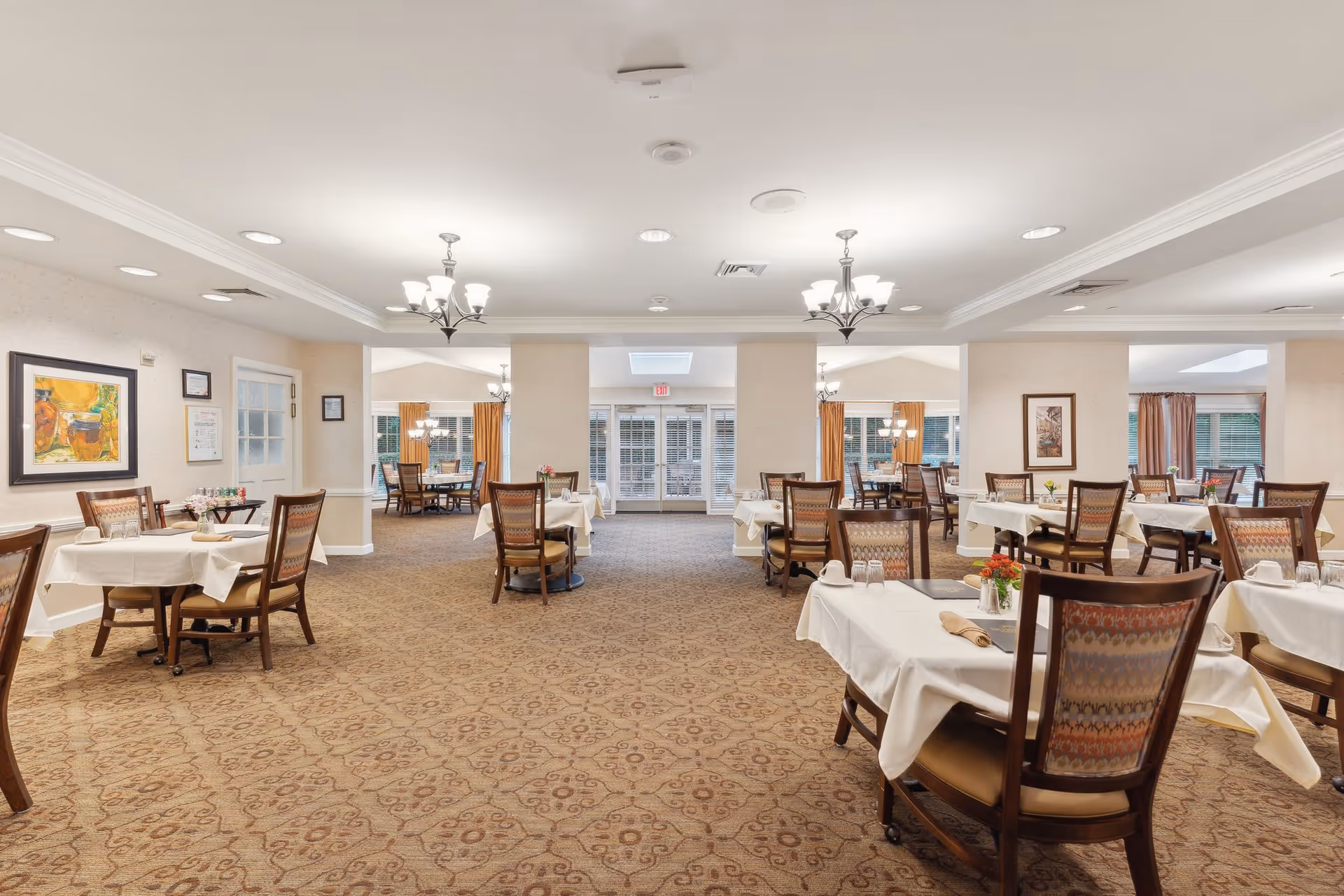 A spacious dining room in an assisted living facility with multiple tables covered in white tablecloths, each set with glasses, napkins, and small flower arrangements. The room has carpeted floors, beige walls, framed artwork, and chandeliers hanging from the ceiling. Large windows and glass doors at the far end allow natural light to enter.