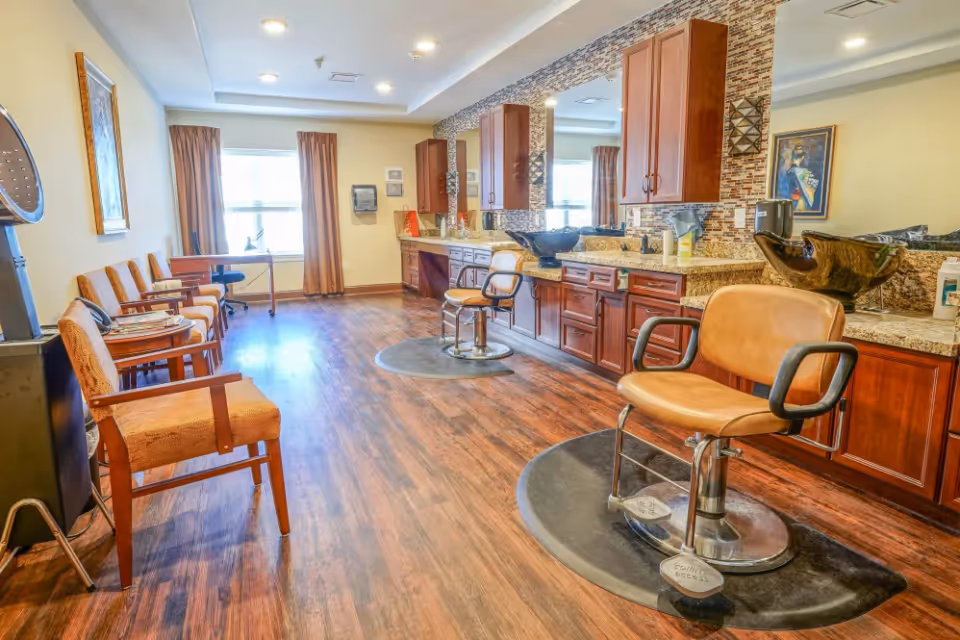 Interior of a salon area in a senior living facility with wooden flooring, two salon chairs on black mats, wooden cabinets with granite countertops, large mirrors, and several cushioned chairs lined up against the opposite wall near a window with brown curtains.