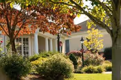 Entrance of a single-story senior living building with white columns, a lamppost, and landscaped gardens framed by trees with autumn leaves.