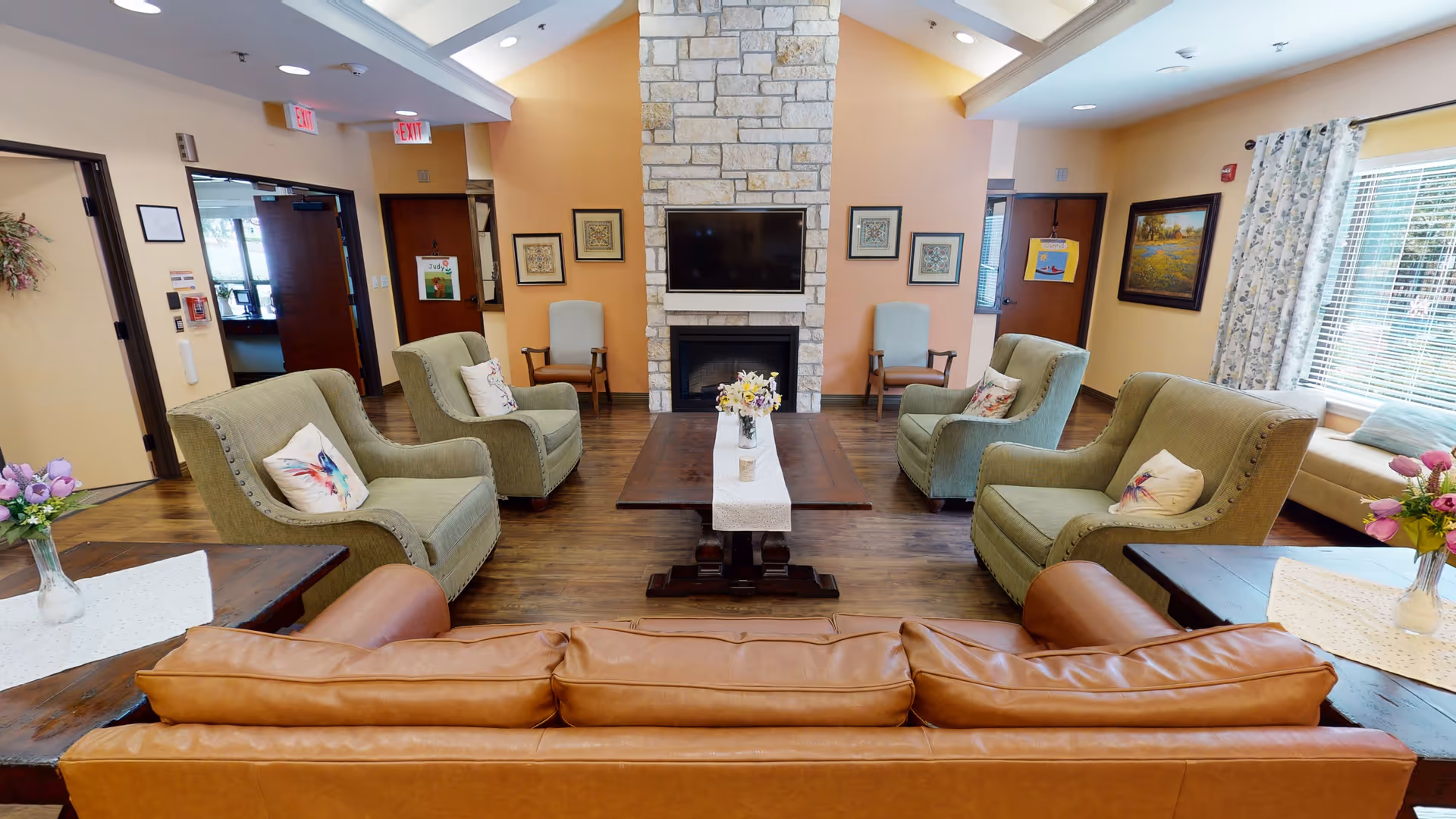 A cozy living room area in Mercy House Living Springs featuring a brown leather sofa, four green armchairs with decorative pillows arranged around a wooden coffee table with a floral centerpiece. The room has a stone fireplace with a mounted flat-screen TV above it, wooden flooring, and large windows with floral curtains letting in natural light.