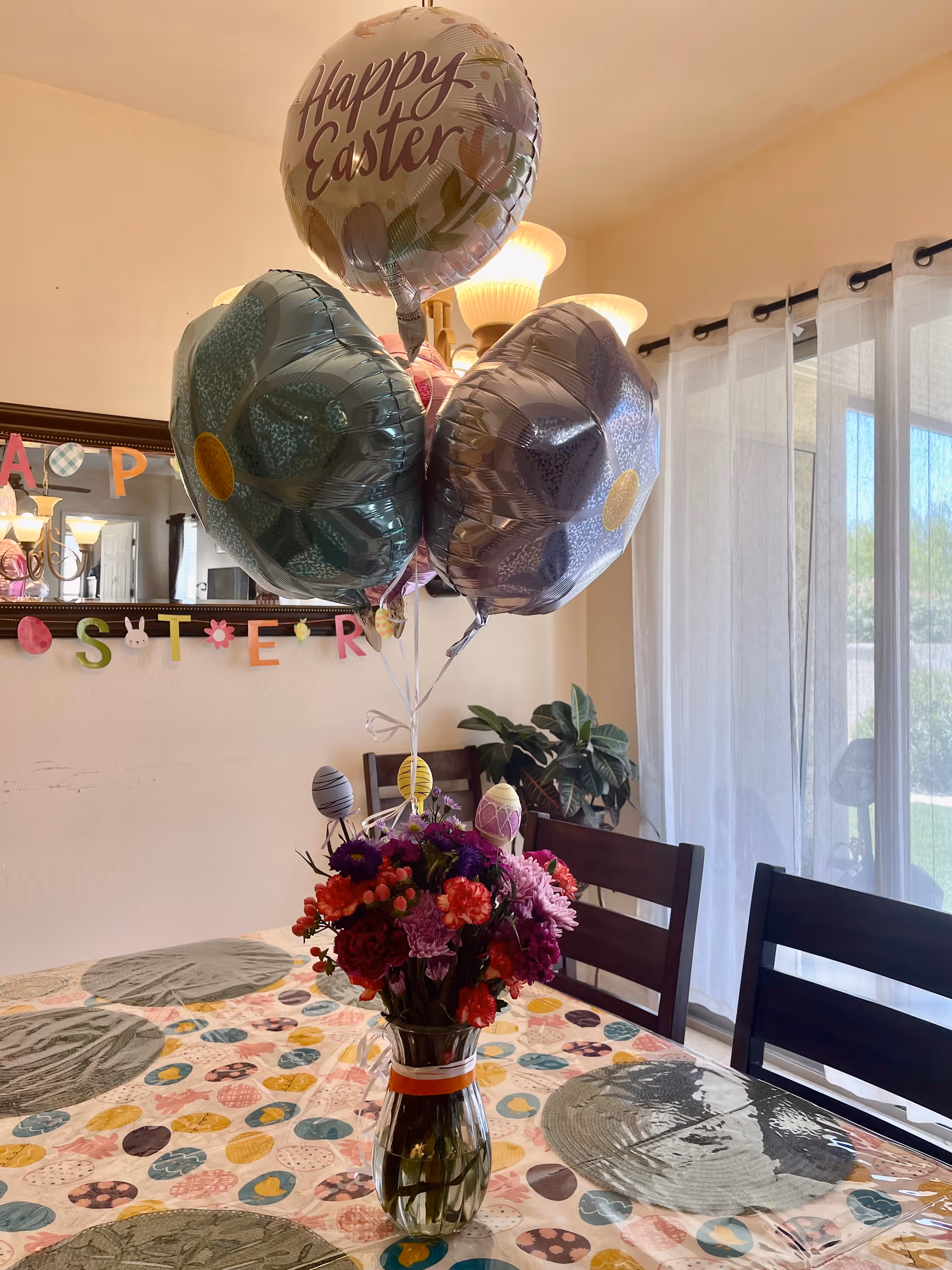 A dining room table decorated for Easter with a colorful tablecloth featuring egg patterns, a vase of vibrant flowers, and three large flower-shaped balloons along with a 'Happy Easter' balloon. In the background, there is a mirror with a festive Easter banner and sheer curtains letting in natural light.
