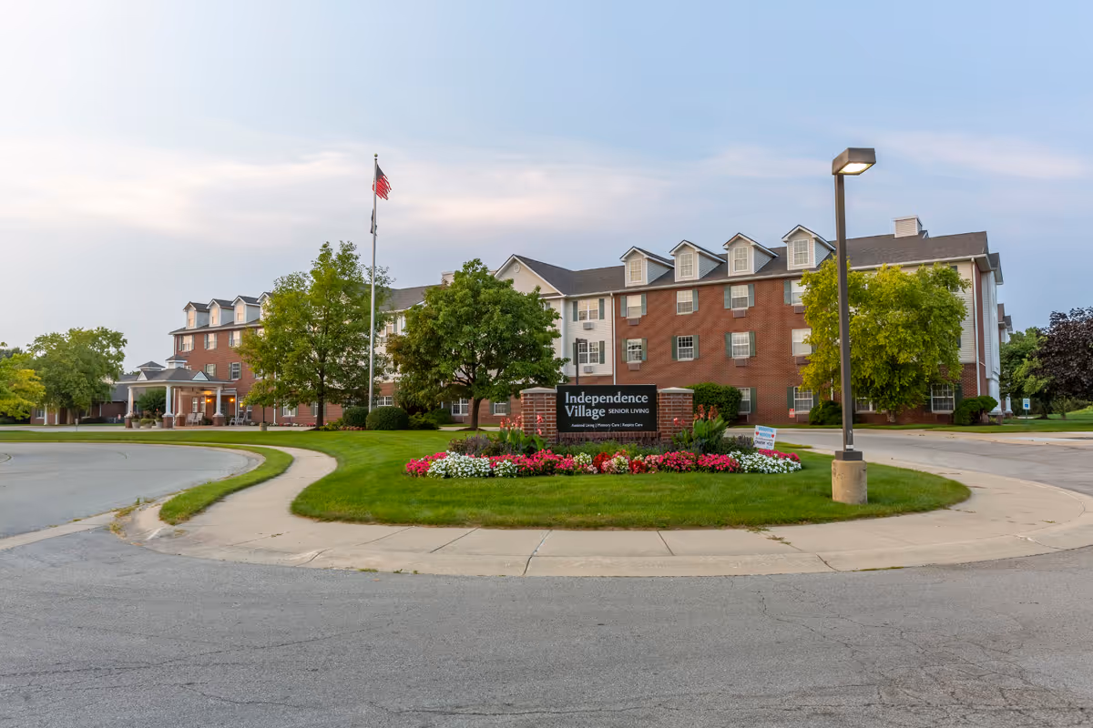Exterior view of Independence Village of Fishers South, a senior living facility with a large brick building, multiple windows, and dormer windows on the roof. The foreground features a circular driveway with a landscaped island containing a sign for the facility, flowers, and trees. An American flag flies on a flagpole near the building, and a streetlamp is visible on the right side.