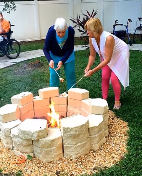 Two women roasting marshmallows over a fire pit made of stacked stone blocks in a grassy outdoor area. One woman has white hair and is wearing a blue top and teal pants, while the other woman has blonde hair and is wearing a white sleeveless top with pink leggings. In the background, there is a person in a wheelchair and some walkers near a white fence.