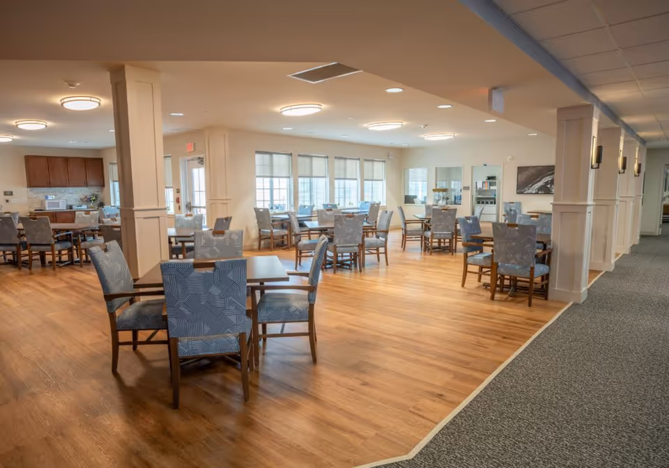 A spacious dining room with multiple wooden tables and cushioned chairs arranged neatly on a wood floor. The room is well-lit with ceiling lights and natural light coming through large windows with blinds. There is a kitchen area with cabinets and a countertop visible in the background. The room has a clean, modern design with light-colored walls and pillars.