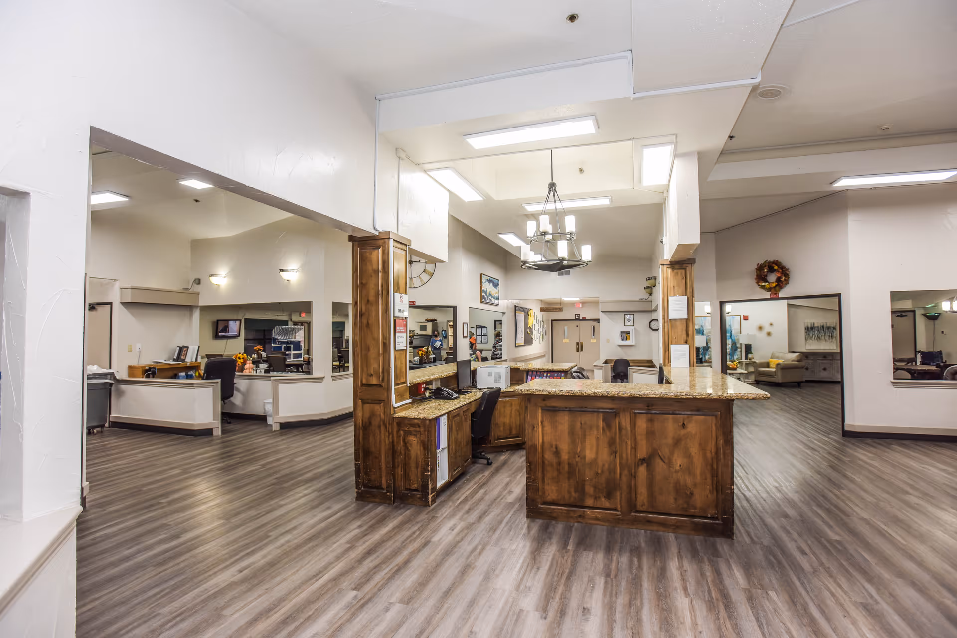 Interior view of a senior living facility reception area with a wooden front desk, chairs, and open spaces leading to other rooms. The floor has wood-like vinyl planks, and the walls are painted white. There are ceiling lights and a chandelier above the desk. Various office and lounge areas are visible in the background.