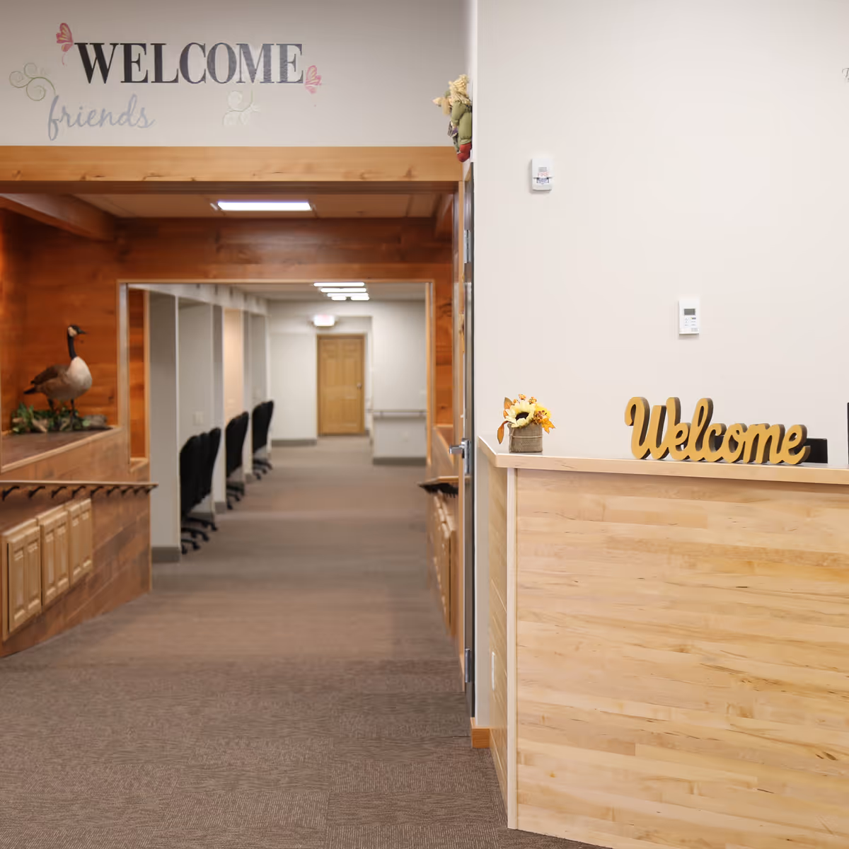 Interior hallway of an assisted living facility with a wooden reception desk on the right displaying a yellow 'Welcome' sign and a small flower arrangement. The hallway has carpeted floors, office chairs lined up along the left wall, and a wooden decorative ledge with a goose figurine. Above the hallway entrance, there is a wall decal that says 'WELCOME friends' with butterfly decorations.