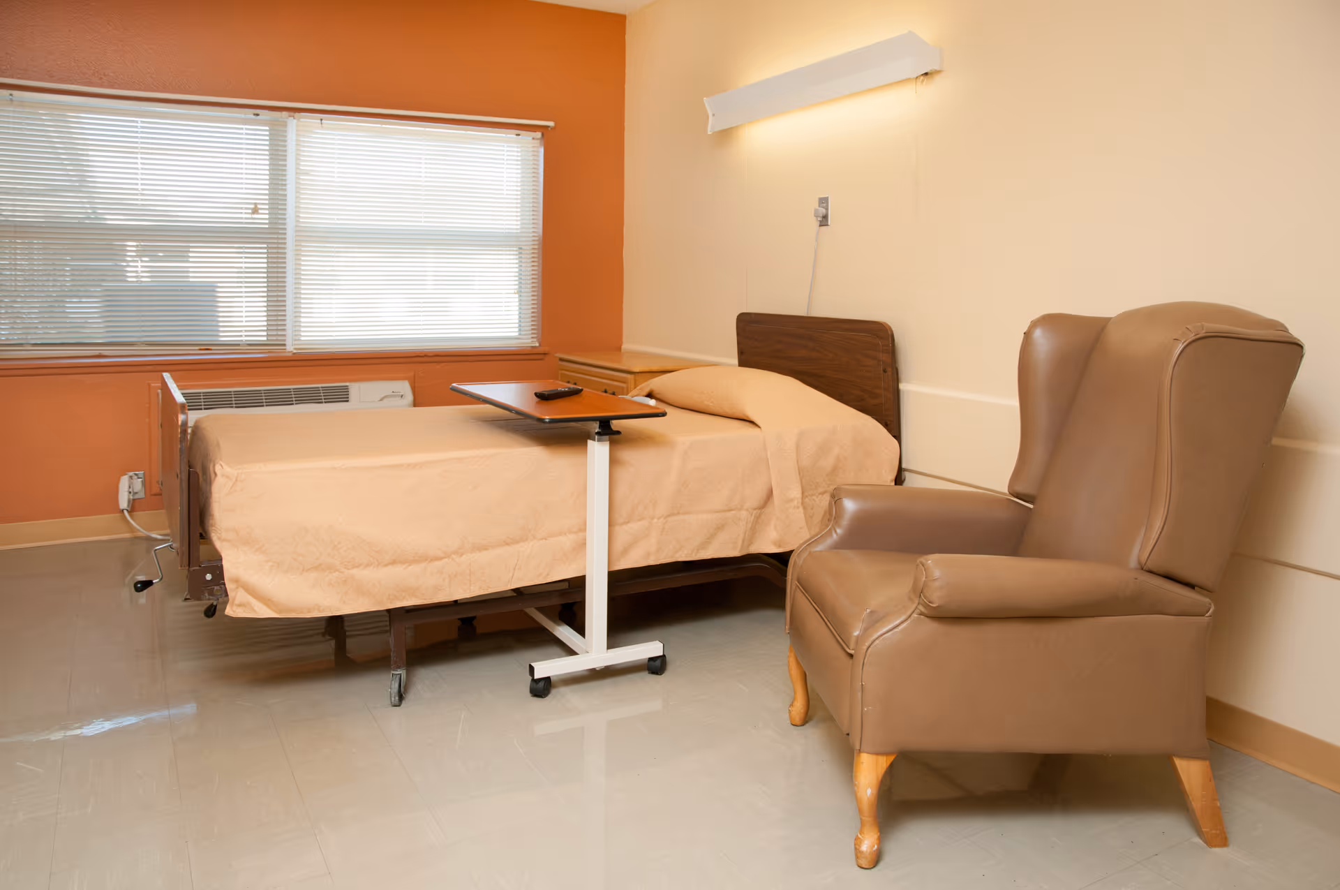 A simple room with a single hospital-style bed covered with a beige blanket, a small overbed table with a remote control on it, a large brown leather armchair, and a window with closed blinds letting in natural light.