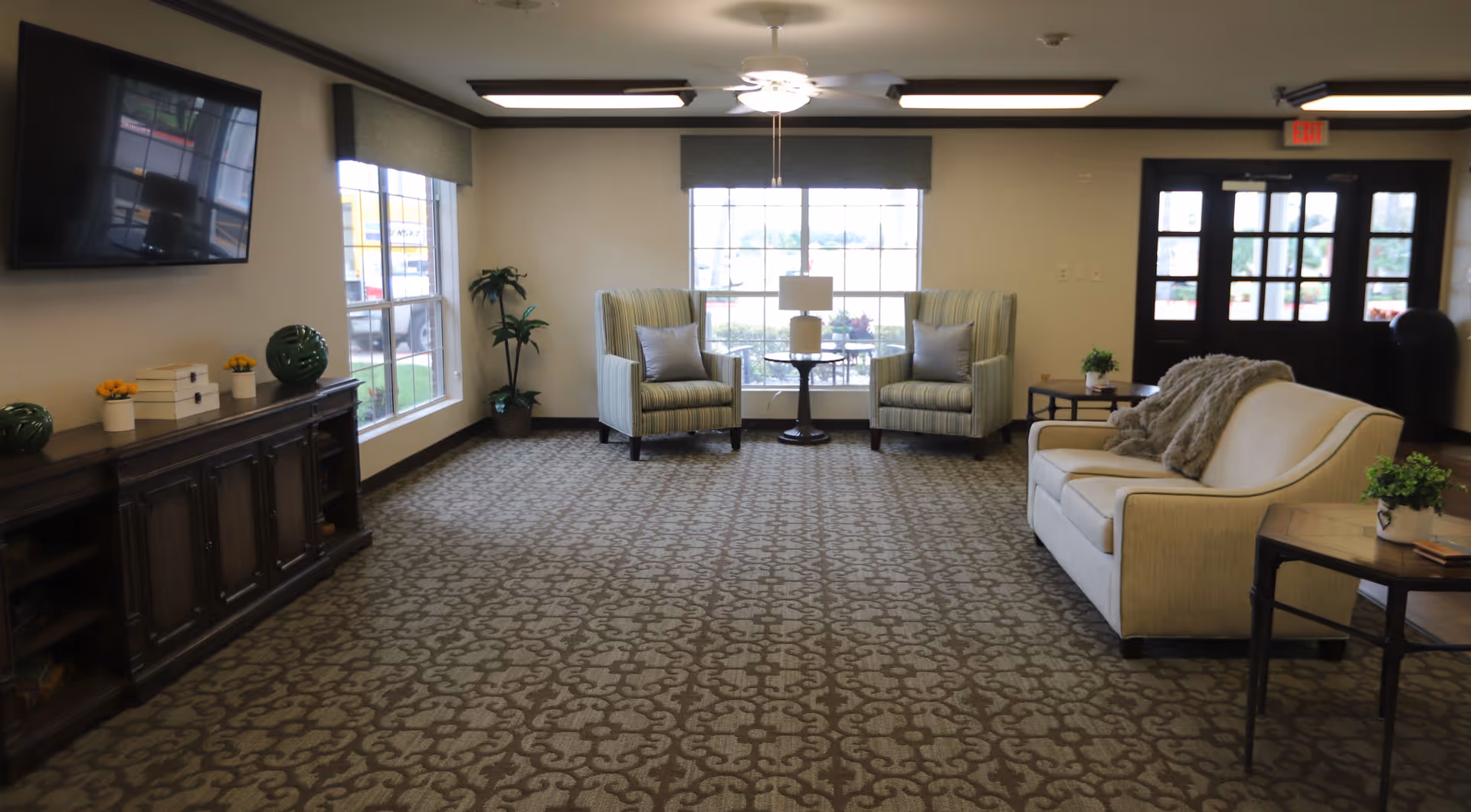 A spacious living room area in Briarcliff Nursing and Rehabilitation Center with patterned carpet, two striped armchairs with pillows, a small round table with a lamp between them, a beige sofa with a throw blanket, side tables with small plants, a large window letting in natural light, a dark wooden cabinet with decorative items, and a wall-mounted flat-screen TV.