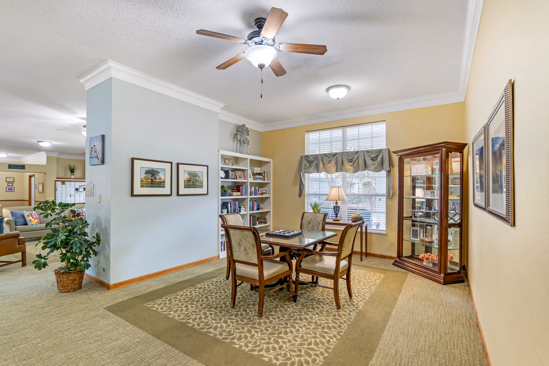 Sunlit common seating area with a wooden table and four chairs on a patterned rug, bookshelves, a display cabinet, and a large window.