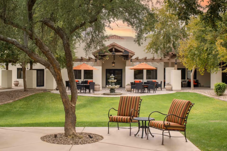 Outdoor seating area at The Village at Ocotillo featuring two striped cushioned chairs with a small table between them, green lawn, trees, and a building with a covered patio, orange umbrellas, and a central water fountain.
