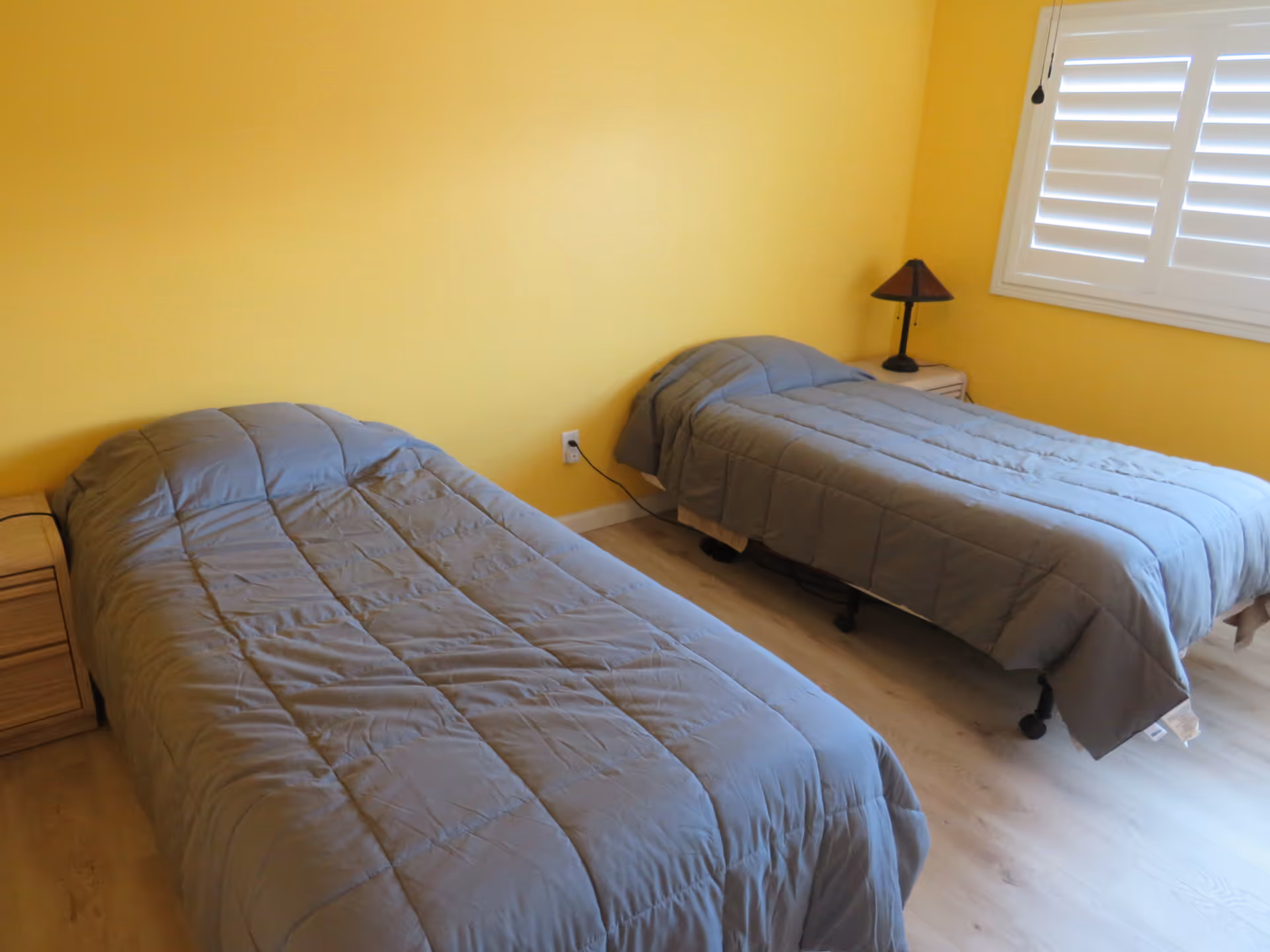 A bedroom with two single beds covered with gray comforters, each bed has a wooden nightstand beside it. One nightstand has a small lamp with a dark shade. The walls are painted yellow and there is a window with white shutters on the right side.