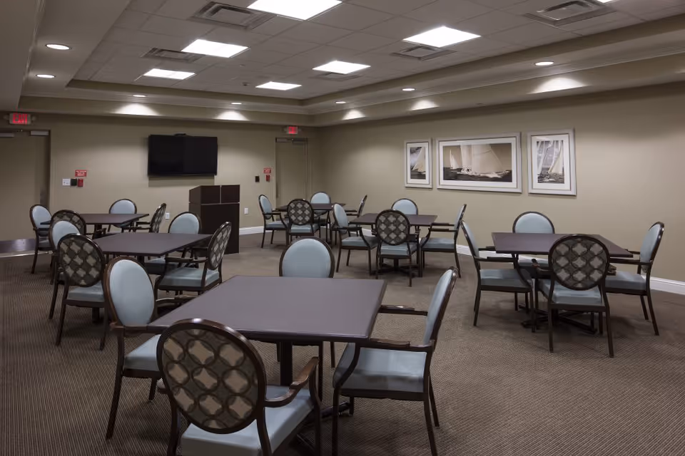Empty communal dining/activity room with multiple tables and chairs, a wall-mounted TV, and framed sailboat pictures on the wall.
