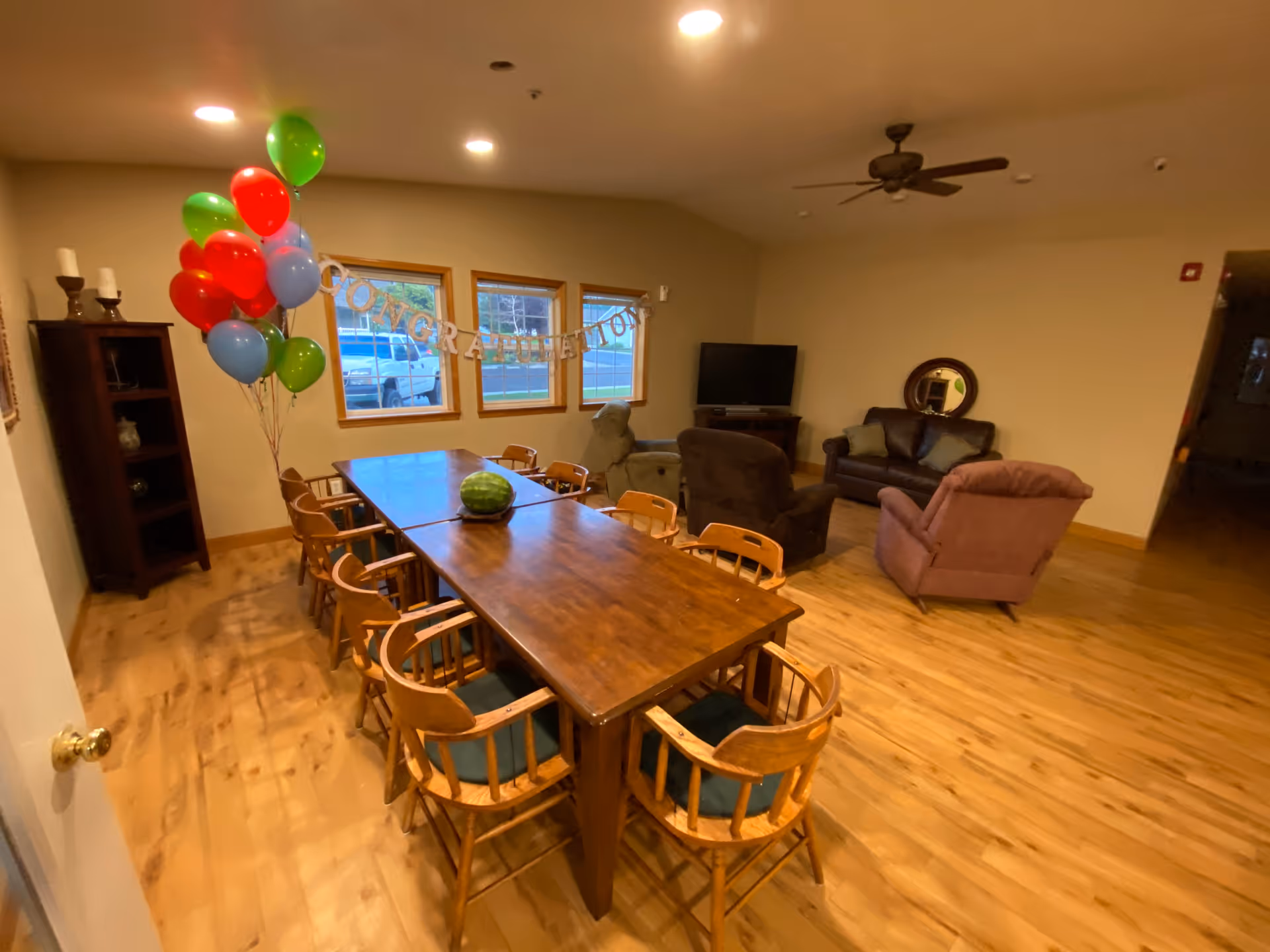 A room with a long wooden table surrounded by wooden chairs, a watermelon placed on the table, colorful balloons tied to the table, and a 'CONGRATULATIONS' banner hanging on the wall. In the background, there are three windows, a TV on a stand, a mirror on the wall, and several upholstered chairs arranged in a seating area.
