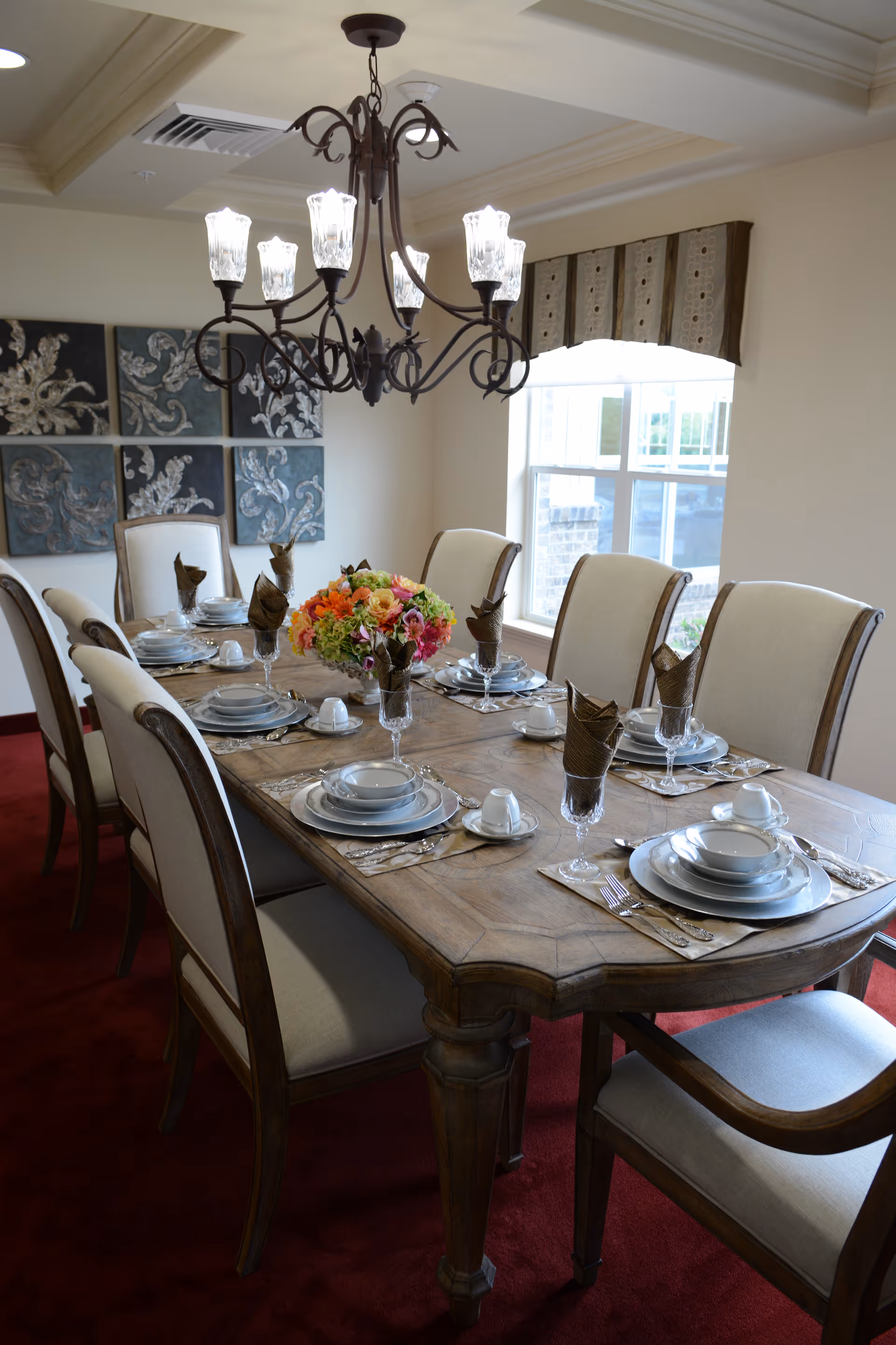 A formal dining room with a wooden dining table set for eight people. The table is adorned with plates, bowls, cups, silverware, and brown folded napkins in glasses. A floral centerpiece is placed in the middle of the table. The room has beige walls, a window with a valance, a chandelier with multiple lights hanging above the table, and decorative wall art in the background.