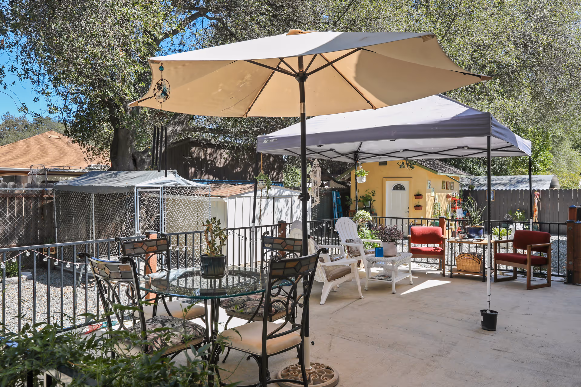 Outdoor patio area with a large beige umbrella over a glass table surrounded by four chairs with patterned cushions. Nearby, there is a canopy tent shading white and red cushioned chairs and a white wicker table with potted plants. The patio is enclosed by a black metal fence, and in the background, there are trees, a wooden fence, and small sheds.