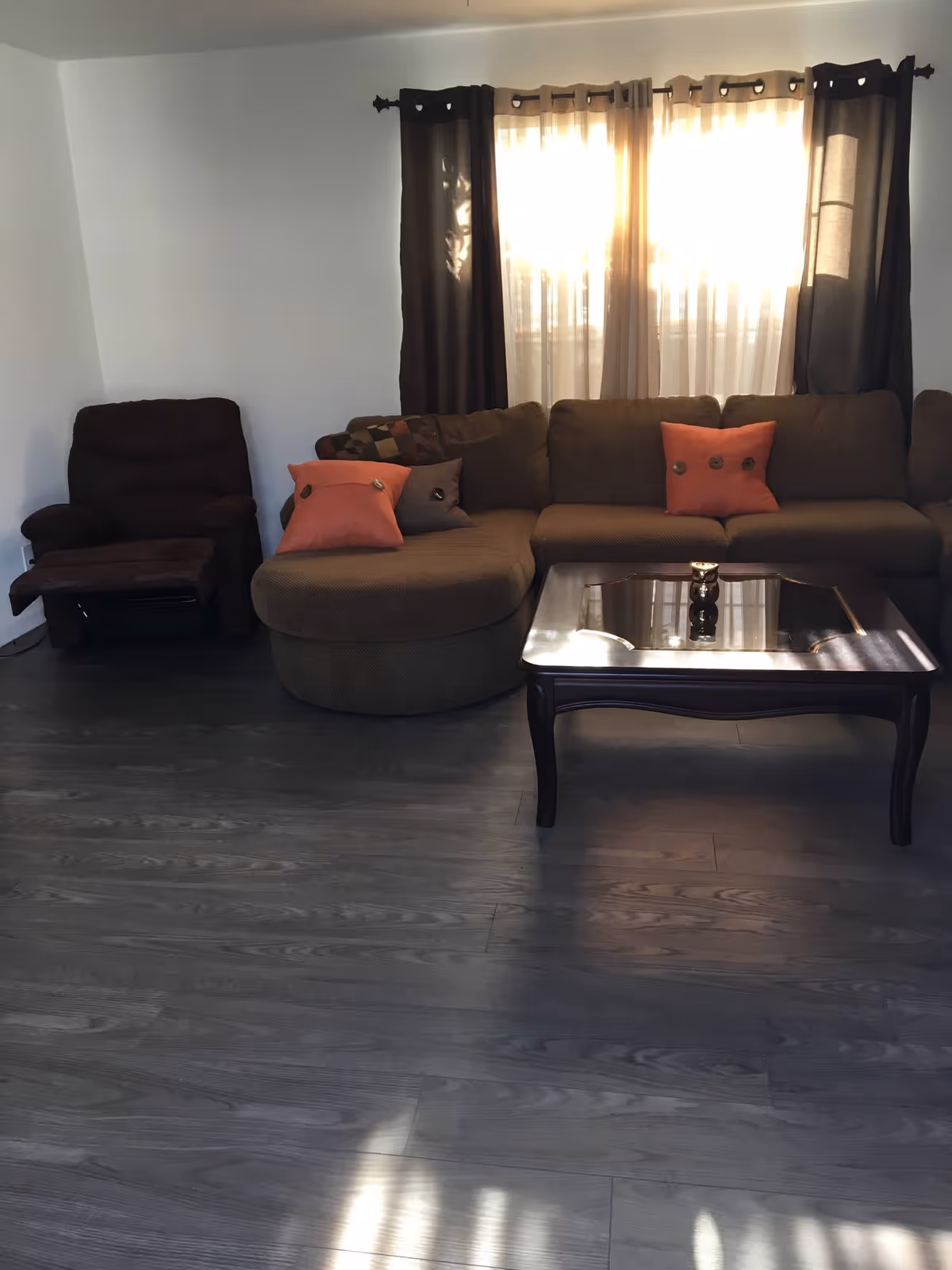 A living room with a brown sectional sofa adorned with orange and brown pillows, a dark brown recliner chair, a wooden coffee table with a glass top, and a window with sheer and dark curtains allowing sunlight to filter through.