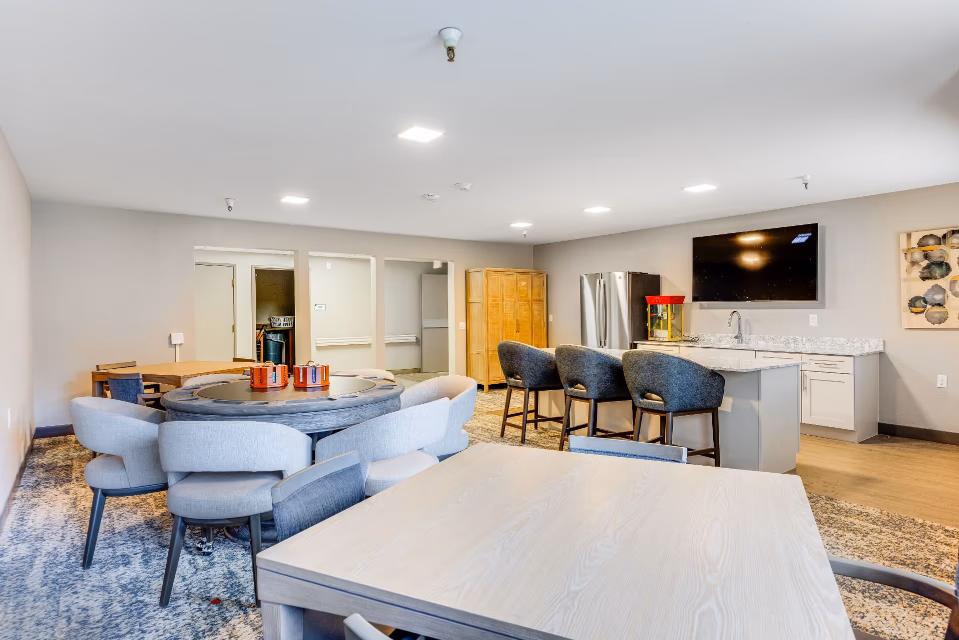 Bright communal lounge and kitchenette with a round dining table, bar seating at an island, wall-mounted TV, and additional tables and chairs.