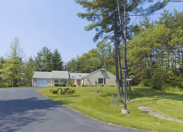 A single-story brick house with a light-colored roof surrounded by a well-maintained lawn and trees. There is a paved driveway leading up to the house, and a lamppost with a small sign is visible on the right side near the edge of the lawn. The background shows a dense area of tall green trees under a clear blue sky.