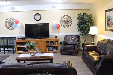 Cozy senior living lounge with leather seating, a TV, bookshelves, and balloons.