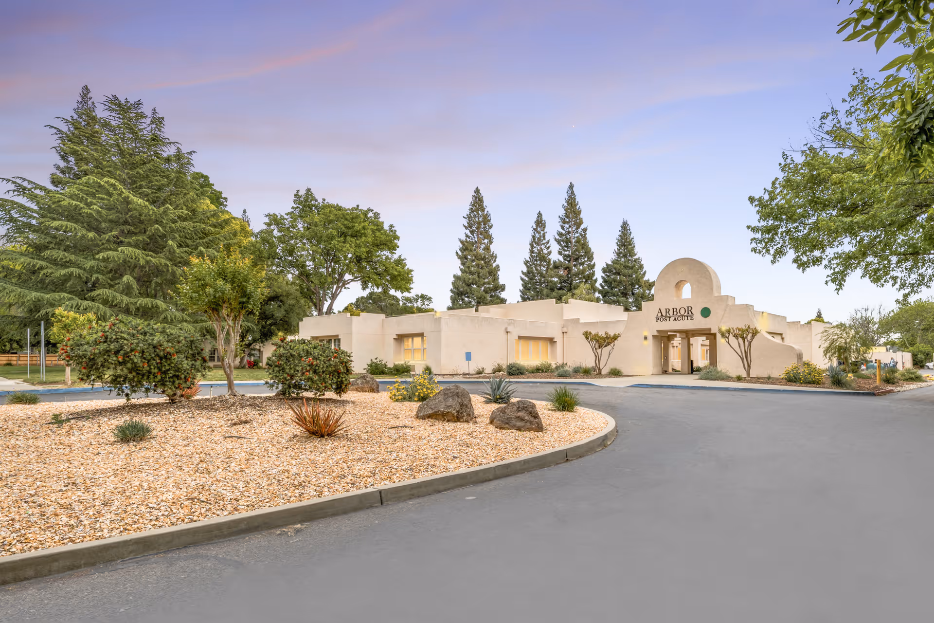 Front exterior of the single-story Arbor Post Acute building with a landscaped circular driveway and trees.