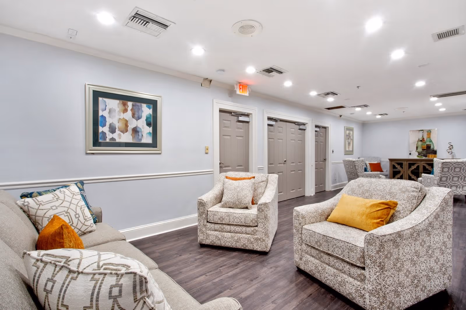 Bright communal lounge with upholstered chairs and sofas, decorative pillows, wall art, and closed doors along the back wall.