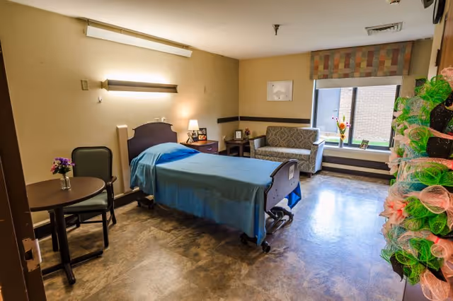 A senior living facility room with a single hospital-style bed covered with a blue blanket, a small round table with two chairs, a nightstand with a lamp and picture frames, a patterned loveseat near a window with a colorful valance, and a decorative wreath hanging on the door.