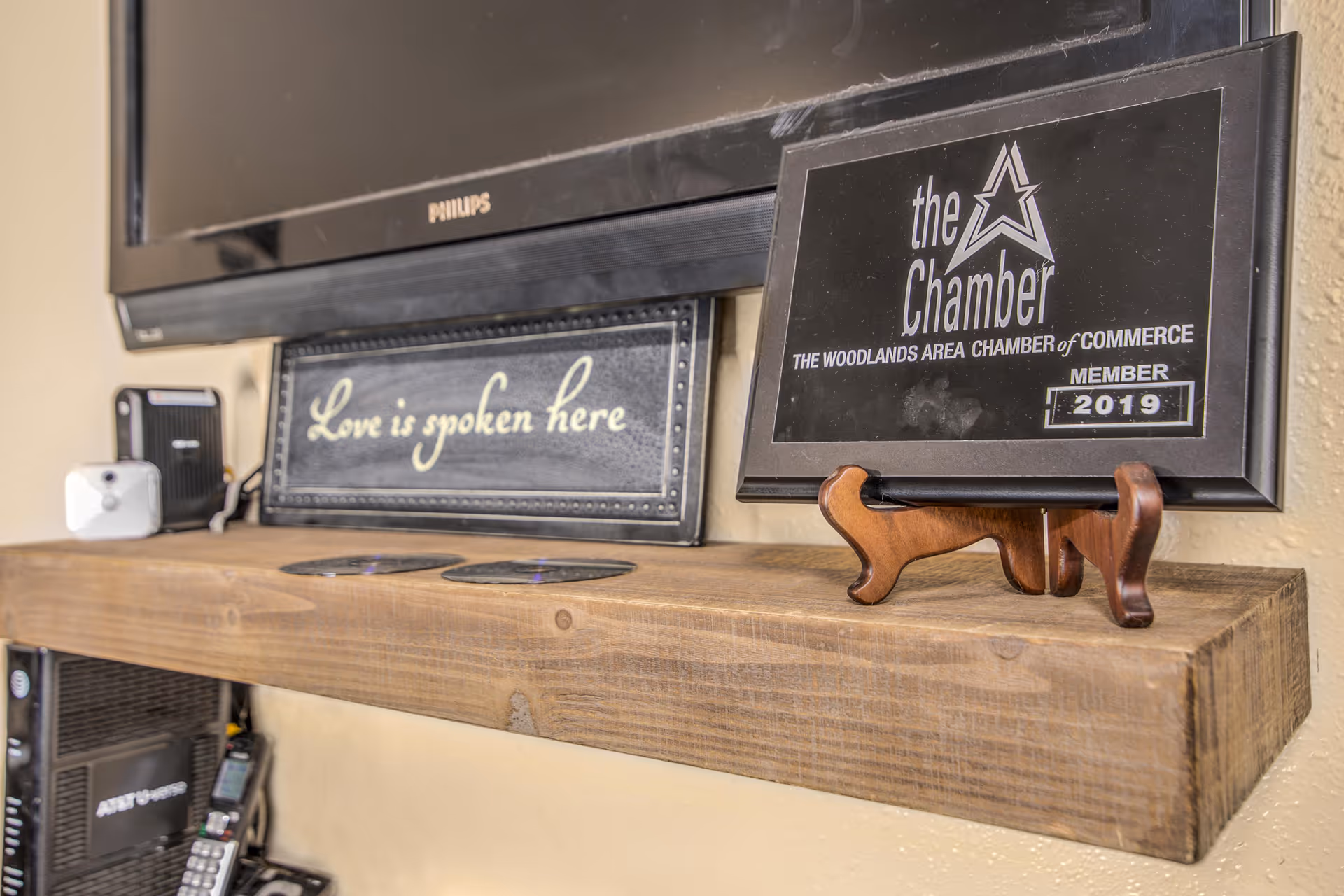 A wooden floating shelf mounted on a beige wall beneath a Philips television. On the shelf, there is a black plaque with the text 'Love is spoken here' and a black framed certificate from The Woodlands Area Chamber of Commerce, member 2019, displayed on a wooden stand. Various electronic devices and cords are visible below the shelf.