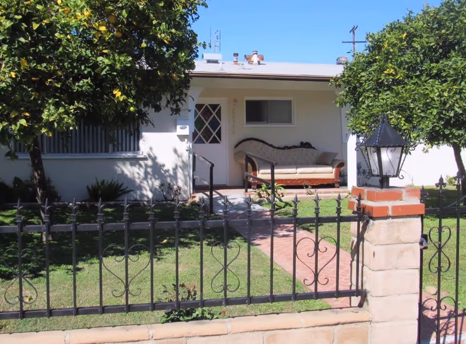 Front exterior of a single-story house with a fenced front yard, brick walkway, and a sofa on the covered porch.