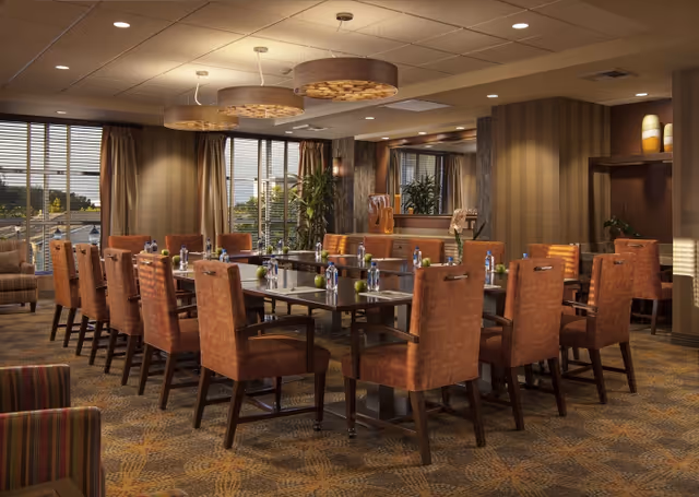 A well-lit dining room with a large rectangular table surrounded by orange upholstered chairs. The table is set with water bottles and green apples. The room features large windows with blinds, patterned carpet, modern ceiling lights, and decorative plants and artwork on the walls.