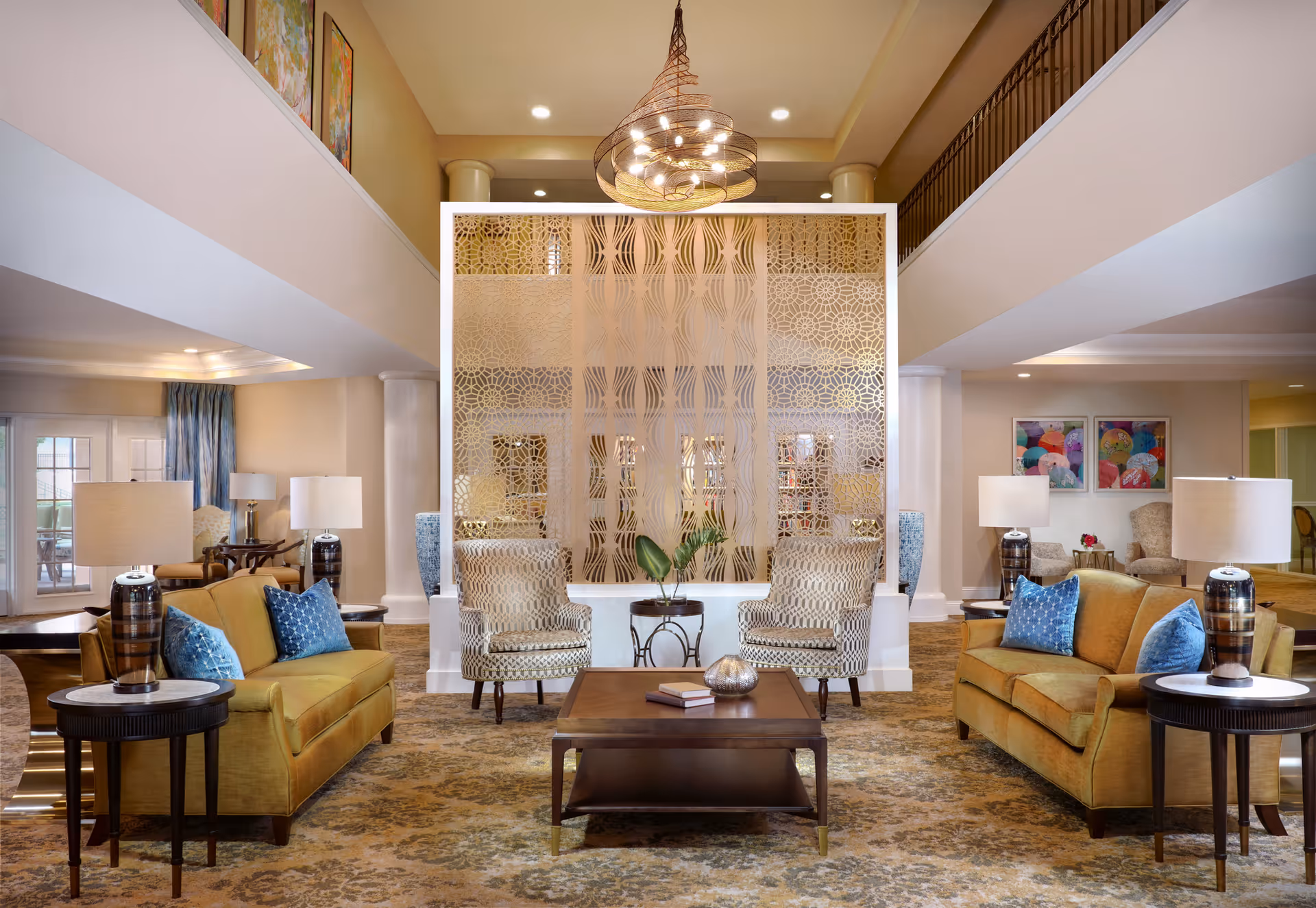 A spacious and elegantly decorated living room area with two mustard yellow sofas, each adorned with blue throw pillows, two patterned armchairs, a wooden coffee table with decorative items, and side tables with lamps. The room features a decorative partition with intricate patterns, a large chandelier hanging from the ceiling, and colorful artwork on the walls.