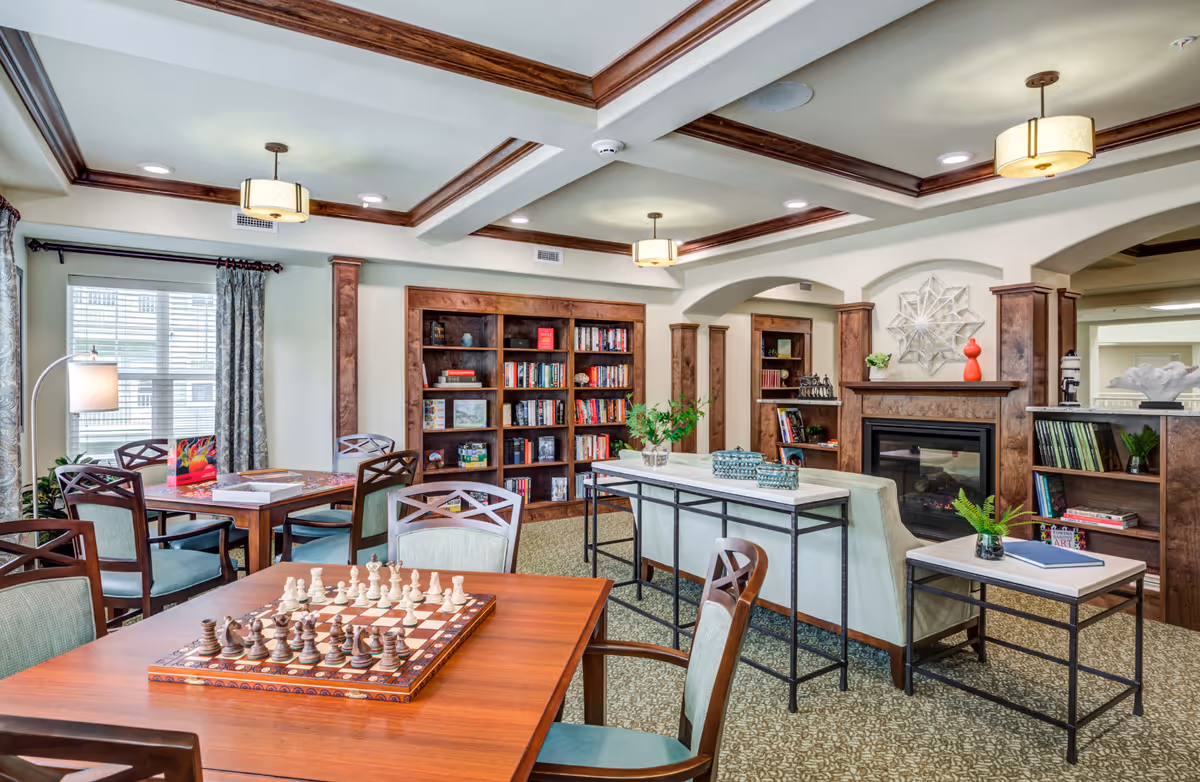 A bright and cozy common area in a retirement living facility featuring a wooden chessboard on a table in the foreground, several chairs, a bookshelf filled with books, a fireplace with decorative items on the mantel, and large windows with curtains allowing natural light to fill the room.