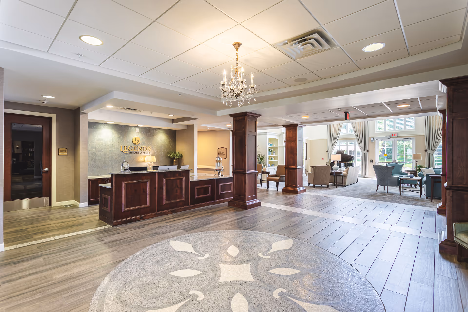 Bright senior living lobby with a wooden reception desk, chandelier, and seating area by large windows.