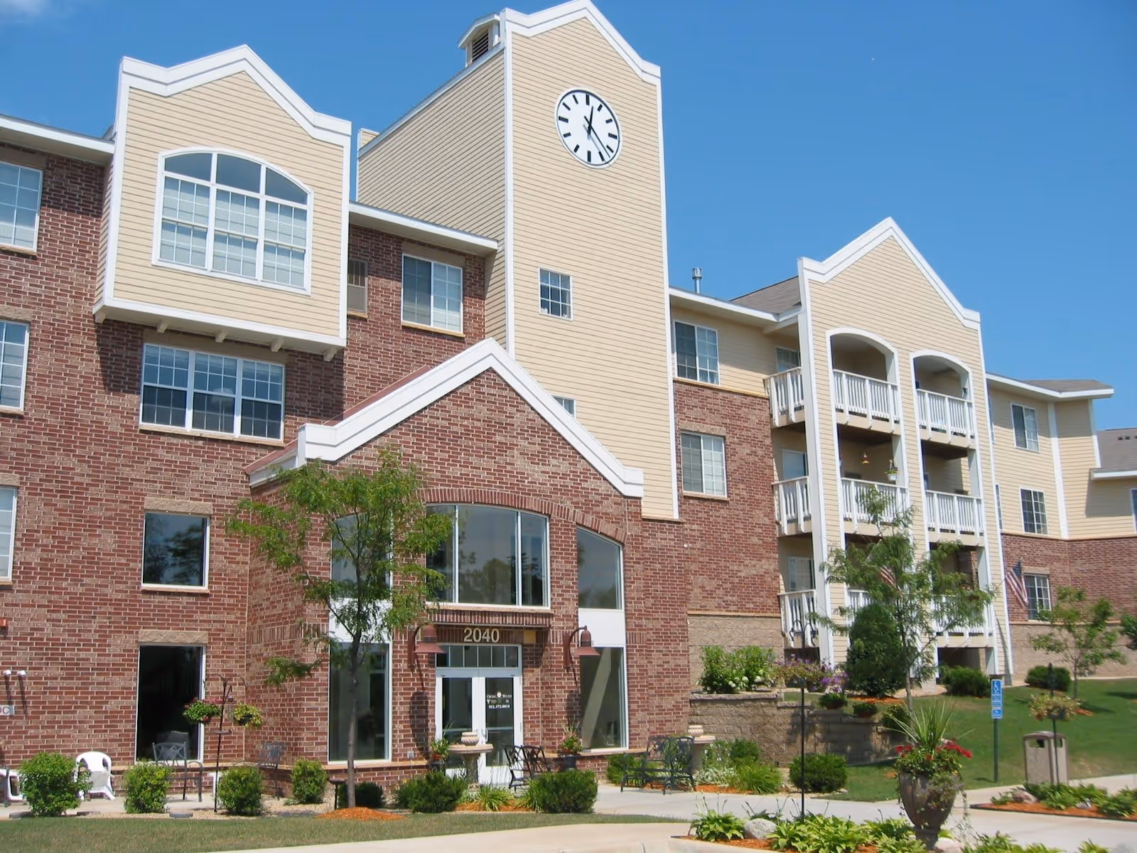 Exterior view of a senior living facility building with a combination of red brick and beige siding. The building has multiple windows, balconies, a clock tower, and a main entrance with the number 2040 above the door. There are small trees, shrubs, and landscaped greenery in front of the building under a clear blue sky.