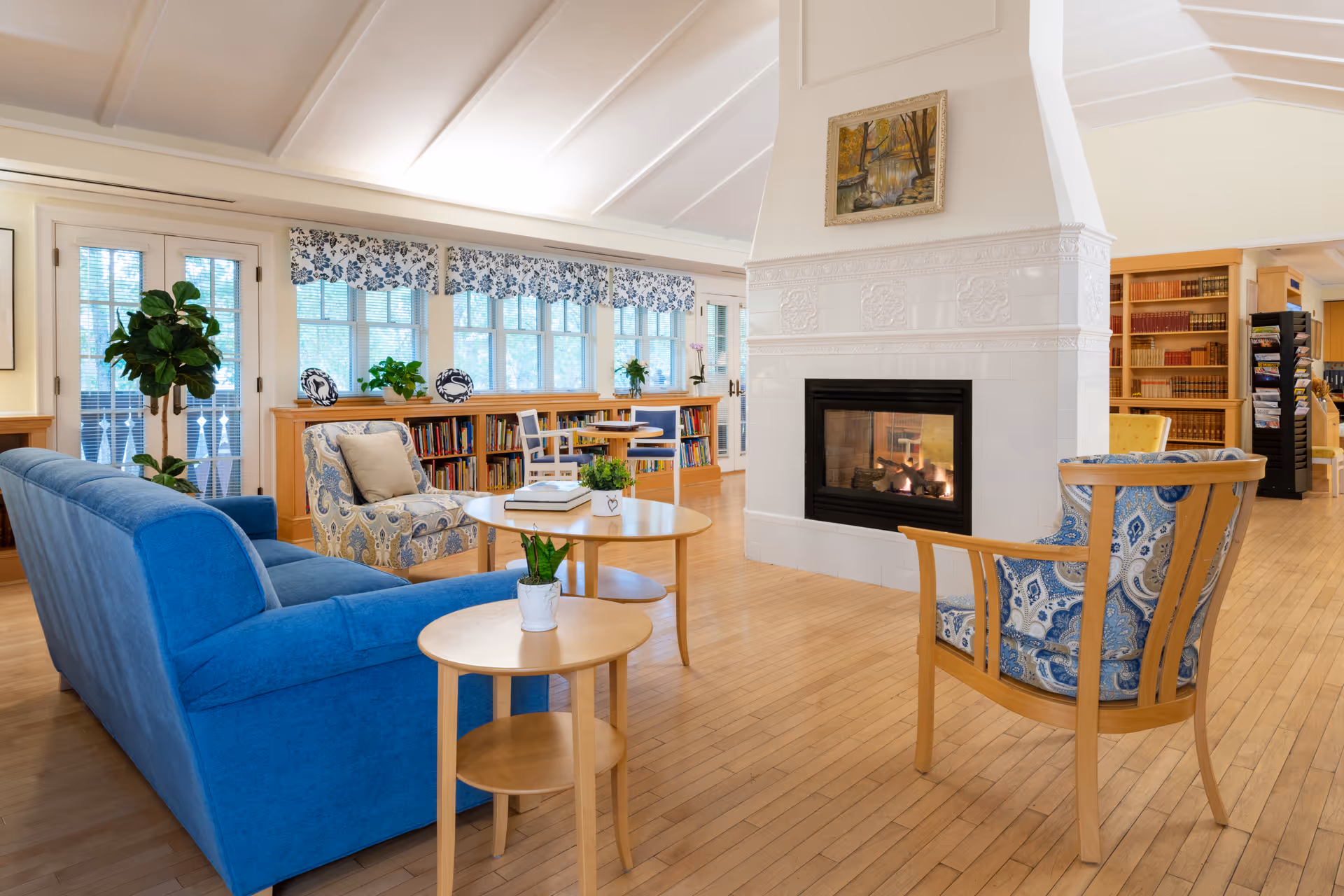 A cozy living room area in a senior living facility with a blue sofa, patterned armchairs, wooden tables with small potted plants, a white tiled fireplace with a painting above it, large windows with floral valances, and bookshelves filled with books in the background.