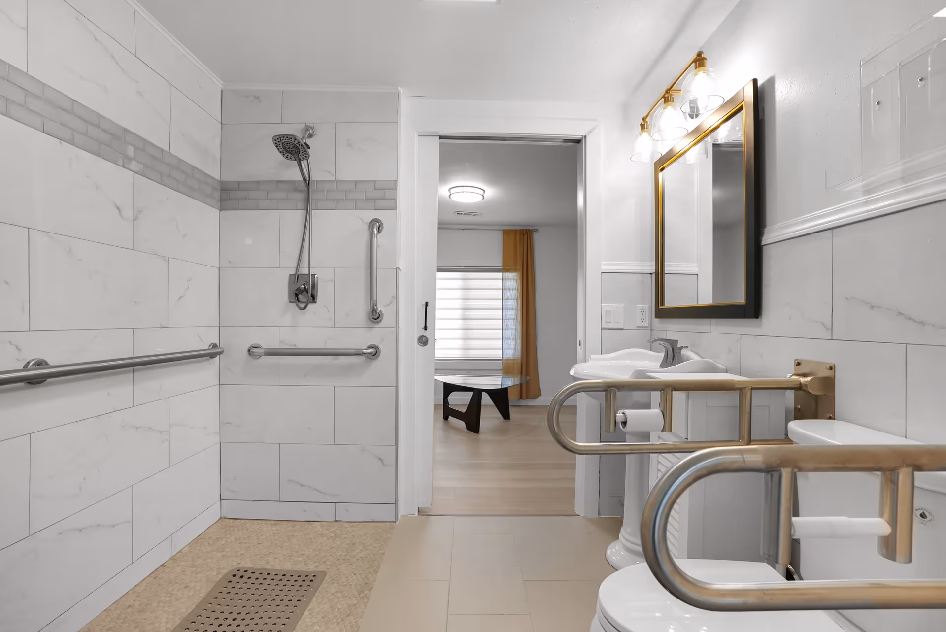 A modern bathroom with white marble tiles and a walk-in shower equipped with stainless steel grab bars. The shower has a handheld showerhead and a tiled floor with a drain. There is a white pedestal sink with a gold-framed mirror and a light fixture above it. The toilet has stainless steel safety rails on both sides. Through an open doorway, a room with a wooden floor, a window with a curtain, and a coffee table is visible.