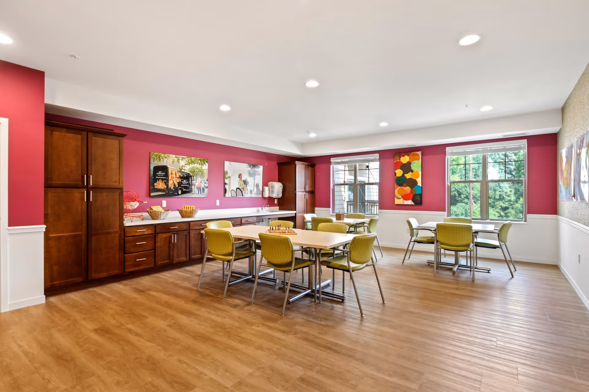 A bright and spacious dining room with wooden flooring, several tables with green chairs, and large windows letting in natural light. The walls are painted with a combination of deep pink and white, decorated with colorful artwork. There are wooden cabinets and a countertop along one wall.