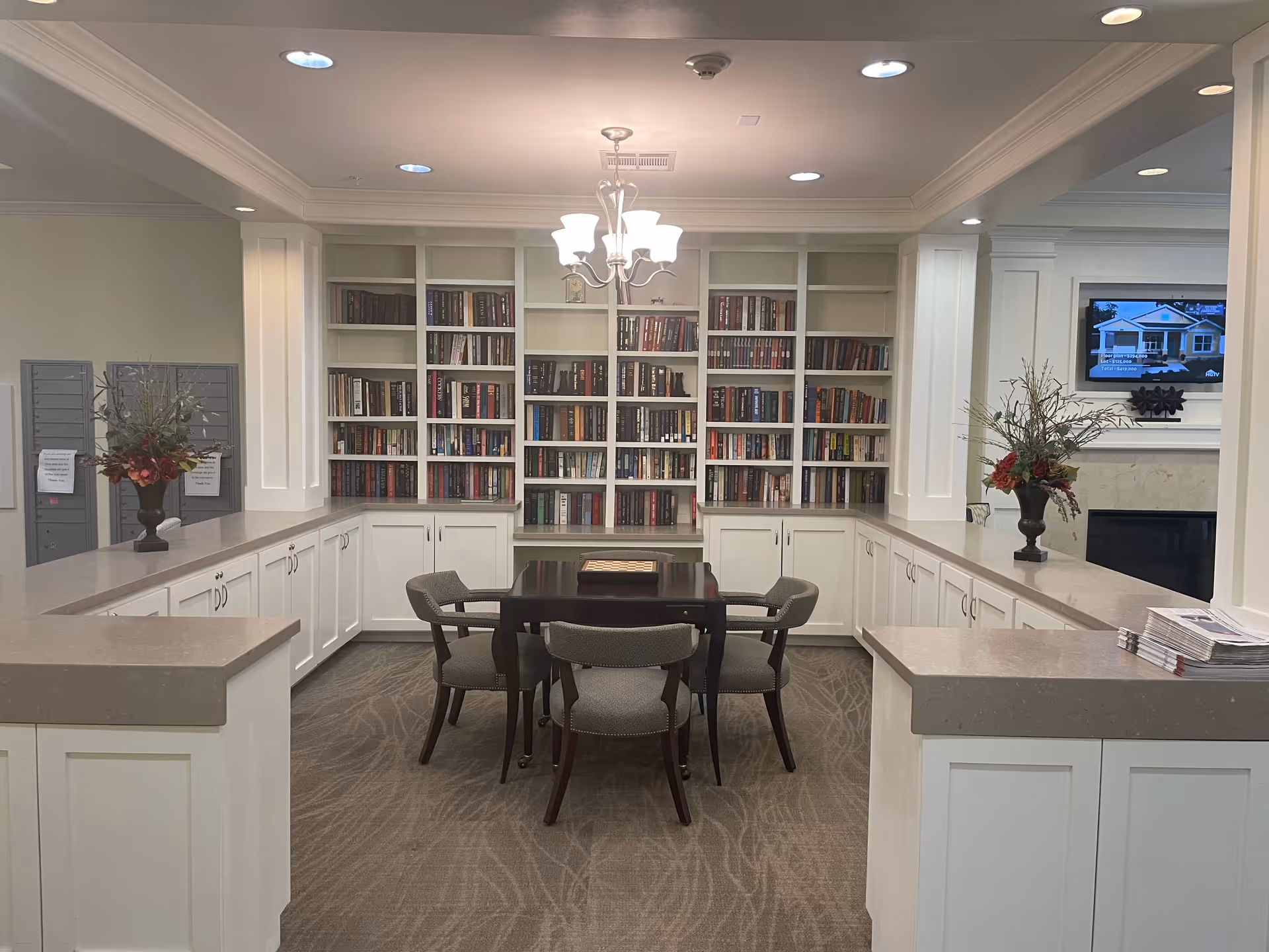 A bright communal library-style room with a central table and chairs, built-in bookshelves and white cabinets, and decorative vases.