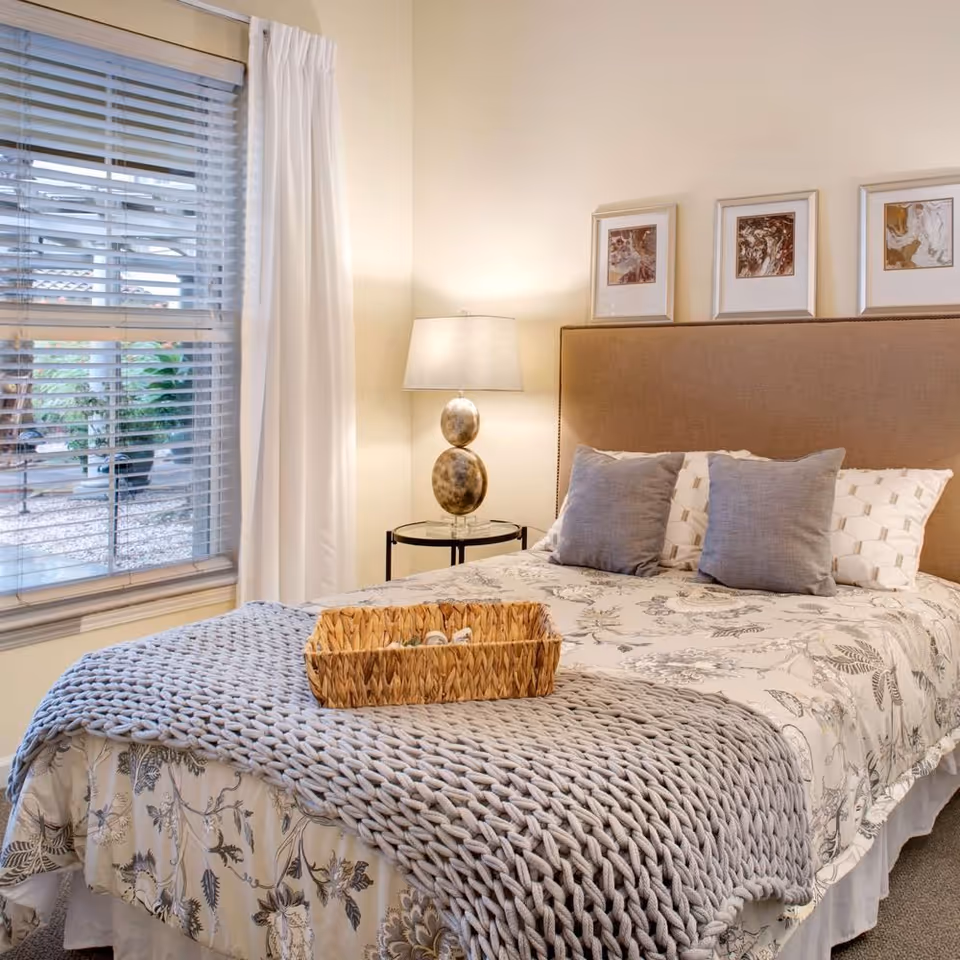 A cozy bedroom with a bed featuring a beige upholstered headboard, floral patterned bedding, two gray pillows, and a chunky knit gray throw blanket. A wicker basket is placed on the bed. Next to the bed is a round glass-top side table with a decorative lamp. Three framed abstract artworks hang on the wall above the headboard. A window with white blinds and curtains is on the left side, showing some greenery outside.