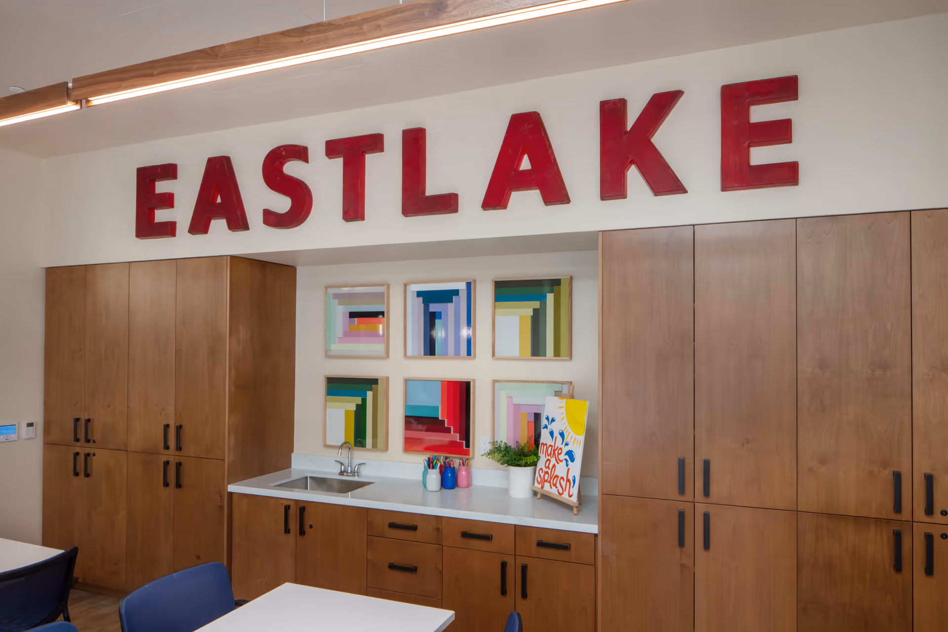 Interior view of a room with wooden cabinets and a countertop with a small sink. Above the cabinets, large red letters spell out EASTLAKE. On the wall above the countertop, there are six colorful framed abstract artworks. On the countertop, there are some paintbrushes in containers, a small potted plant, and a sign that reads 'make a splash'. Several blue chairs and white tables are partially visible in the foreground.