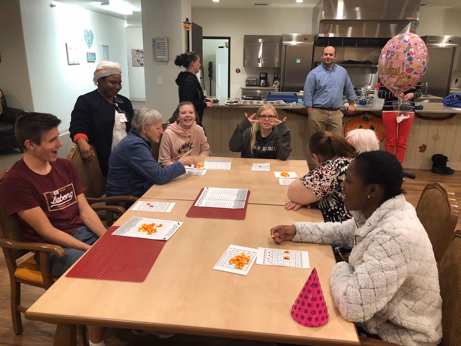 A group of people, including elderly individuals and younger people, sitting around a table playing a game with candy corn pieces on bingo cards in a well-lit room. In the background, a man stands near a kitchen counter, and a person holds a pink birthday balloon. The setting appears to be a communal area in an assisted living facility.