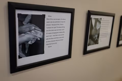 Two framed black and white photographs with accompanying text are hung on a beige wall. The closest frame shows a photo of hands holding each other and a story about a person named Ginny. The second frame is slightly blurred and also contains a photo and text.