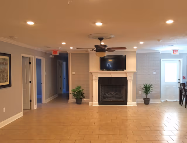 Interior view of a common area with a fireplace and a mounted flat-screen TV above it. The room has beige tiled flooring, light gray walls, and recessed ceiling lights. There are two potted plants on either side of the fireplace, a ceiling fan with lights, and an exit door to the right. A hallway with open doors is visible on the left side of the image.