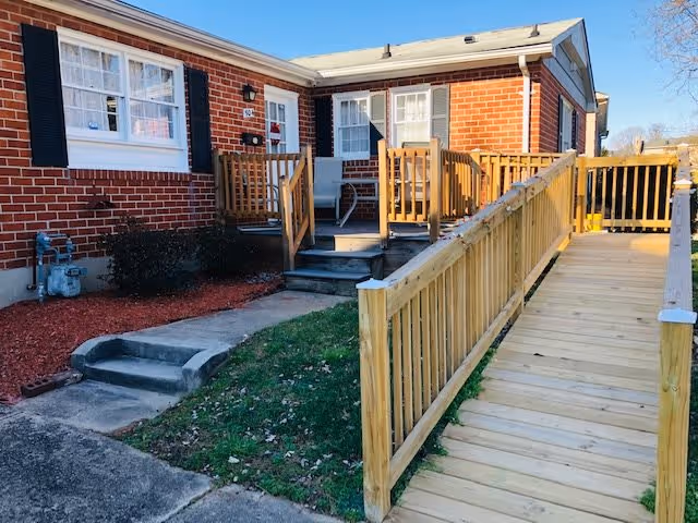 Exterior view of a single-story brick building with white-framed windows and black shutters. There is a wooden wheelchair ramp with railings leading up to the entrance, alongside a small set of stairs. The area around the building has some grass, mulch, and a concrete walkway.